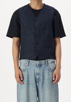 Mann trägt eine dunkelblaue, geknöpfte Weste über einem schwarzen kurzärmeligen Hemd und hellblaue Jeans mit lockerem Sitz, steht vor einem einfarbigen Hintergrund.