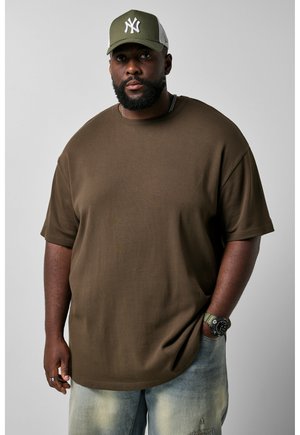 Man met baard die een olijfgroene baseballpet draagt, een oversized bruin T-shirt, lichte spijkerbroek, horloge en ring, staand tegen een effen achtergrond.