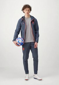 Athlete in a dark gray tracksuit with orange accents holds a blue and red soccer ball. Features a zippered jacket and slim-fit pants.