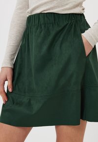 Jupe en daim vert avec taille élastique, poches latérales et texture lisse. Présente un design simple et une silhouette droite.
