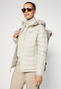 Lys beige puffer-jakke med kviltet mønster, glidelåslommer og hette, kombinert med en beige topp og bukser. Brune solbriller fullfører antrekket.