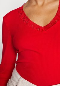 Punainen ribbineuleinen pitkähiha, jossa on V-mallinen kaula-aukko ja kukkakangasta. Kangas vaikuttaa pehmeältä, ja muotoilu on vartaloonmyötäinen.