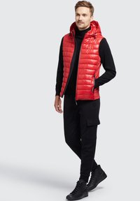 Rød puffervest med hætte, der har et quiltet design og lynlåslommer foran, båret over en sort turtleneck og sorte cargo-bukser.