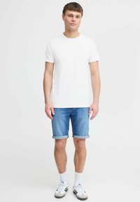 Ung mand iført en ensfarvet hvid t-shirt, opsmøgede blå denimshorts, hvide sokker og hvide Adidas sneakers med sorte striber, stående.
