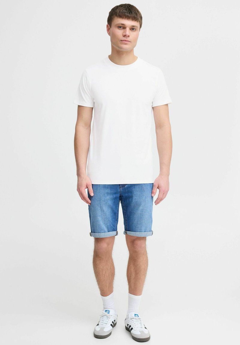 Ung mand iført en ensfarvet hvid t-shirt, opsmøgede blå denimshorts, hvide sokker og hvide Adidas sneakers med sorte striber, stående.