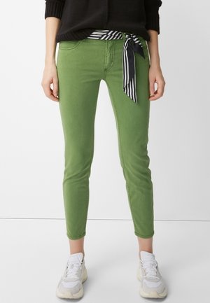Kvinde iført grønne slim-fit bukser med et sort-hvidt stribet stofbælte, hvide sneakers og en sort sweater.
