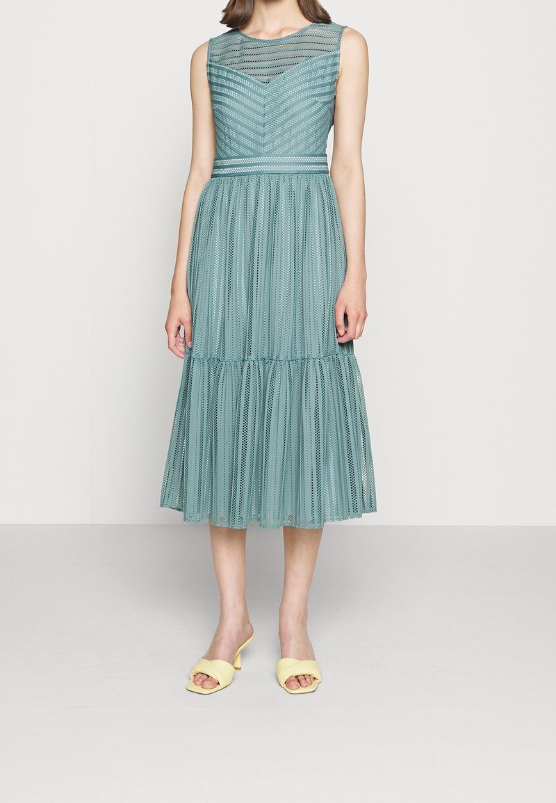 Robe midi sans manches couleur sarcelle en tissu rayé transparent avec une taille cintrée, portée avec des sandales à talons ouvertes jaune clair.
