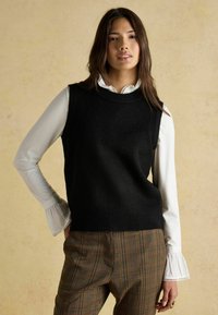 Musthave mustriga särk ilma varrukateta ümmarguse kaeluse ja valge pikavarrukalise särgi, millel on volangkäised. Riietuse täiendavad pruunid ruudulised püksid.