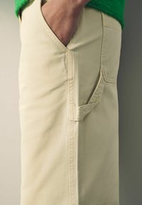 Beige puuvillahousut, joissa on sivutasku, sileä pinta ja kapea malli. Näkyvät ompeleet saumoissa.