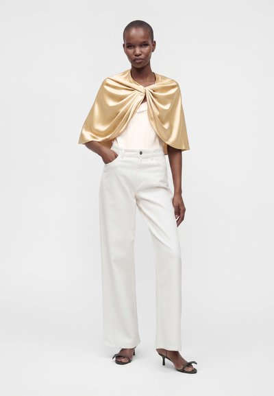 Femme debout portant un châle en satin doré sur un haut blanc, un pantalon blanc taille haute à jambes larges et des talons ouverts noirs sur un fond uni.