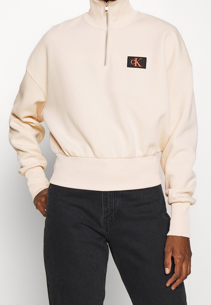 Jasnopomarańczowa bluza z zamkiem błyskawicznym, z prążkowanymi mankietami i krawędzią, z czarną naszywką z logo "CK". Miękka tkanina, luźny krój, minimalistyczny design.