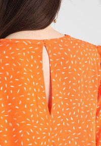 Orange bluse med en lille knaplukning i ryggen, med en nøglehulåbning og et letvægtsstof prydet med hvide abstrakte mønstre.
