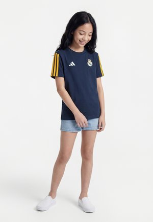 Fille souriante, portant un maillot bleu marine du Real Madrid avec des rayures jaunes, un short en denim clair et des chaussures blanches sans lacets, debout devant un fond blanc.