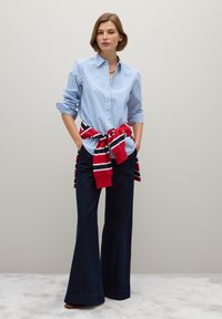 Chemise à rayures bleues et blanches avec un col boutonné, associée à un pantalon évasé sombre. Un pull à rayures rouges noué à la taille ajoute un élément de contraste.
