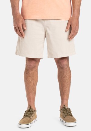 Mand iført beige kordfløjls-shorts, en ferskenfarvet t-shirt og beige snøresneakers, stående mod en ensfarvet baggrund.