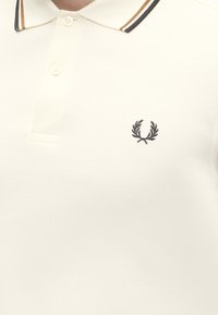 Polo crème avec patte de boutonnage à deux boutons, bords de col rayés et petit logo couronne de laurier noir sur la poitrine gauche.