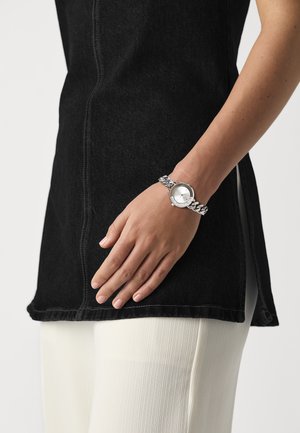 Montre bracelet en argent avec un cadran rond, bandoulière à maillons, portée sur une main. La personne qui la porte a la peau olivâtre et est habillée de vêtements noirs et crème.