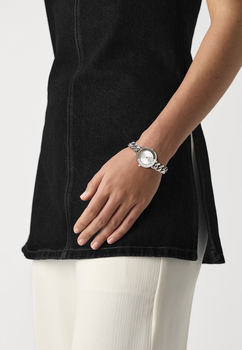 Silver bracelet watch with a round face, linked chain band, worn on a hand. The wearer has olive skin and is dressed in black and cream clothing.