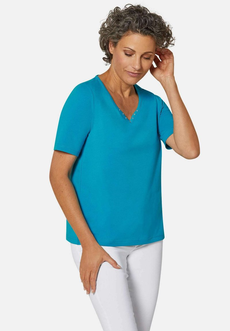 Turquoise V-hals T-shirt met korte mouwen, met een zachte textuur en drie decoratieve studs bij de hals. Gekleed met witte broek.