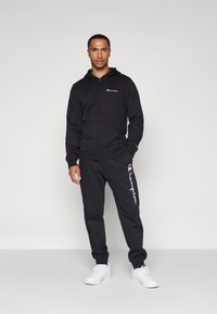 Schwarze Zip-Hoodie mit Fronttaschen und Logo, kombiniert mit passenden Jogginghosen mit seitlichem Logo. Getragen mit weißen Sneakers.