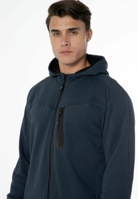 Haut de survêtement zippé bleu marine avec une capuche noire. Présente une poche zippée diagonale et une texture lisse avec une conception ajustée.
