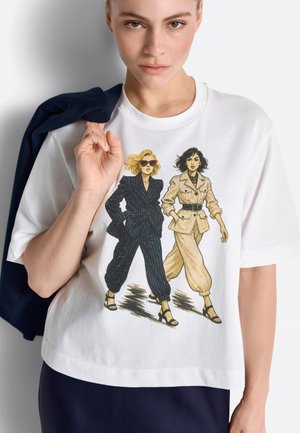 Donna con maglietta bianca con illustrazione di due donne sicure di sé in completi eleganti e sandali che camminano in avanti.