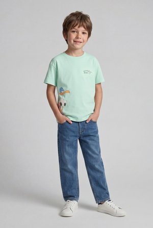 Niño joven sonriendo con las manos en los bolsillos, con una camiseta verde menta con un pájaro de dibujos animados y vaqueros azules sobre un fondo liso.