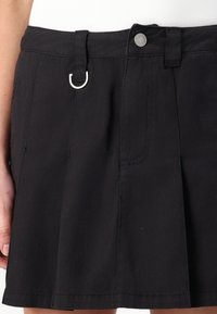 Čierna skladaná sukňa vyrobená z odolnej tkaniny. Obsahuje detail s predným okom a zapínanie na gombík s dvojitými bočnými vreckami.