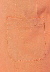 Tissu orange avec une texture lisse présentant une poche rectangulaire. Des coutures visibles bordent la poche, ajoutant un détail au design.
