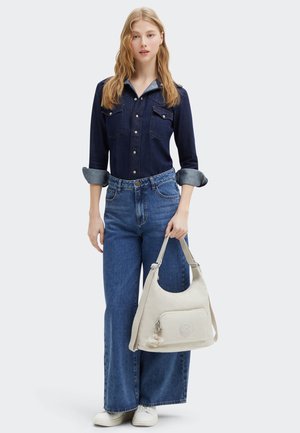 Lys beige skuldertaske med et afrundet design, frontlomme med lynlås og en nøglering som accent. Sammen med en mørk denimskjorte og brede blå jeans.