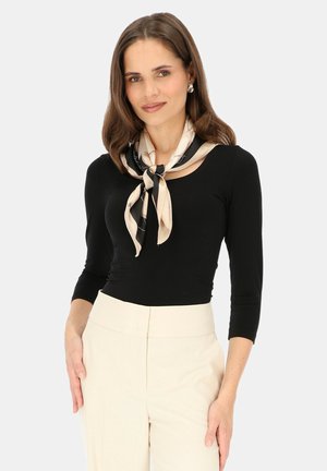 Kobieta o brązowych włosach długości do ramion, ubrana w czarną bluzkę z długim rękawem, kremowe spodnie z wysokim stanem oraz jasnobeżowy szalik w czarny wzór zawiązany na szyi.