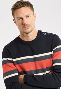 Homme aux cheveux courts portant un pull marine à rayures horizontales rouges, blanches et grises, avec des boutons à l'épaule, regardant l'appareil photo les bras croisés.