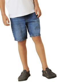 Shorts en denim d'un bleu moyen, avec une coupe classique, des ourlets repliés et un design à cinq poches. Portés avec une chemise blanche et des chaussures grises.