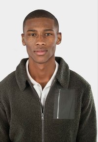 Giacca in fleece verde scuro con zip frontale, colletto a punta e tasca sul petto. Presenta un tessuto testurizzato e una zip liscia come dettaglio.