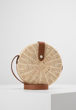 Ronde geweven rotan crossbodytas met bruine leren band en sluiting, geplaatst op een witte ondergrond tegen een effen achtergrond.