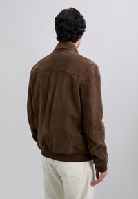 Mies, jolla on tummat kiharat hiukset, pukeutuneena ruskeaan mokkatakkiin ja valkoisiin housuihin, nähtynä takaa yksinkertaista taustaa vasten.