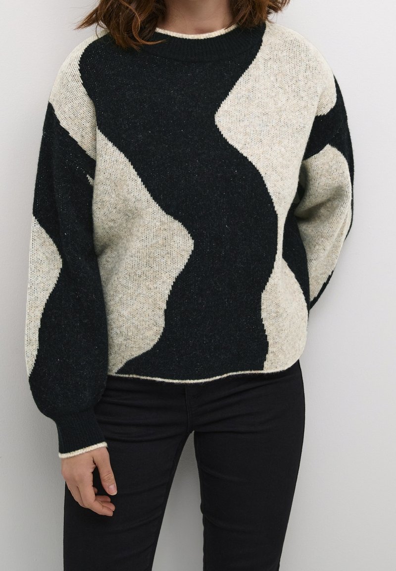 Personne portant un pull oversize noir et beige avec un motif ondulé et des manches ballon, assorti à un pantalon noir, sur un fond uni.