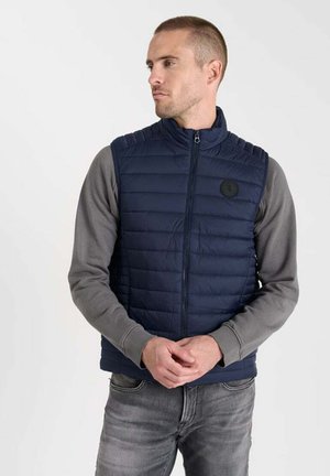 Marineblaues Steppweste mit gepolstertem Textur und hohem Kragen. Mit einem Frontreißverschluss und einem runden schwarzen Logo-Patch ausgestattet. Trägt man über einem grauen Langarmshirt.