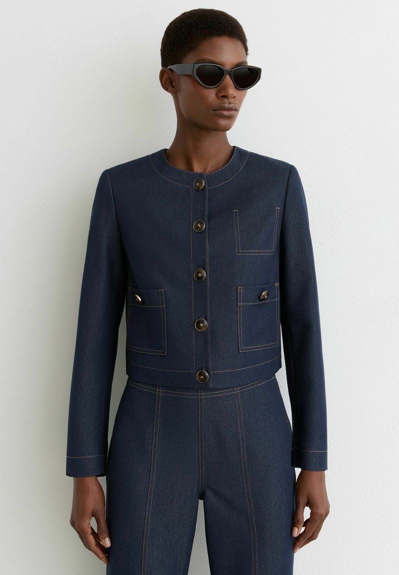 Donkere denim cropped jack met een ronde hals, knopen in goudkleur, twee voorzakken en contrasterende gele stiksel details.