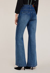 Dżinsy o szerokich nogawkach i wysokim stanie w średnim niebieskim kolorze, wykonane z gładkiego denimu. Posiadają tylne kieszenie oraz brązową skórzaną łatę na pasie.