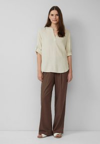 Hellbeige Leinenhemd mit V-Ausschnitt und umgeschlagenen Manschetten, kombiniert mit lockeren braunen Hosen und weißen Sandalen. Glatte Textur, lässige Passform.