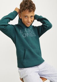 Felpa verde con tasca frontale e tessuto texturizzato. Presenta la scritta "JACK & JONES" stampata in grigio chiaro. Abbinata a pantaloni chiari a righe.