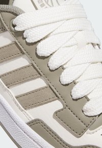 Vit sneaker med grå accenter, featuring texturerad mocka och släta syntetiska material. Tjocka vita snören och framträdande tre-randig design.