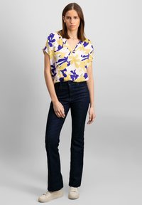 Bluse med blomstermotiv i gul, lilla og hvid med V-udskæring, parret med mørkeblå lige bukser og lyse sneakers.