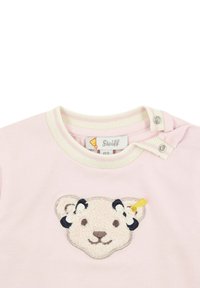 Helles rosa Baumwoll-T-Shirt mit einem strukturierten, gestickten Bärenkopf, schwarzen Akzenten und einem gelben Band sowie Druckknopfverschluss an der Schulter.
