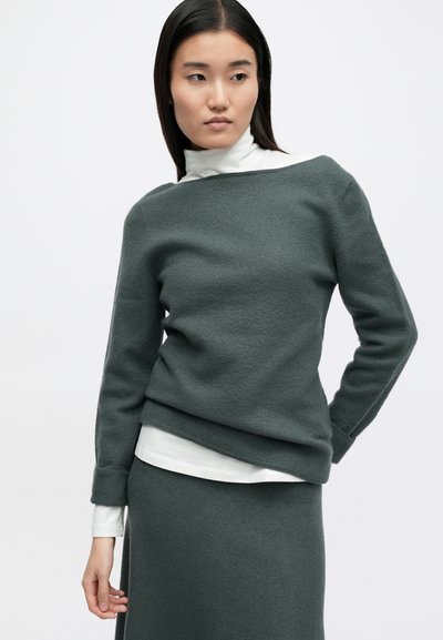 Γκρι, υφασμένος σκούφος με φαρδιά λαιμόκοψη, συνδυασμένος με λευκό πουλόβερ τύπου λαιμόκοψη από κάτω. Το σύνολο διαθέτει μακριά μανίκια και χαλαρή εφαρμογή.