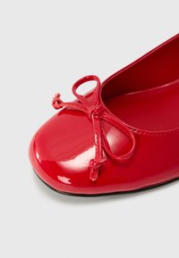 Röd lackskinnsballetsko med rund tå, prydd med en dekorativ rosett av matchande glänsande material. Slät yta, svart sula.