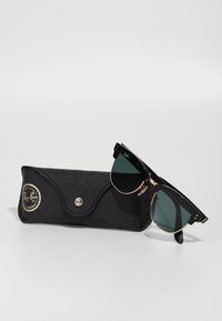 Musta Ray-Ban aurinkolasit tummanvihreillä linssillä, kultaisilla yksityiskohdilla ja mustilla varsilla; mukana musta nahkainen kotelo logolla.