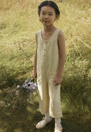 Tuta in lino giallo chiaro con design senza maniche, bottoni frontali e due tasche anteriori; bambino che tiene un mazzo di fiori.
