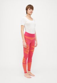 Fitted pink leggings with orange geometric patterns, ribbed ankle cuffs, and a high waistband. Worn with a plain white short-sleeve top.